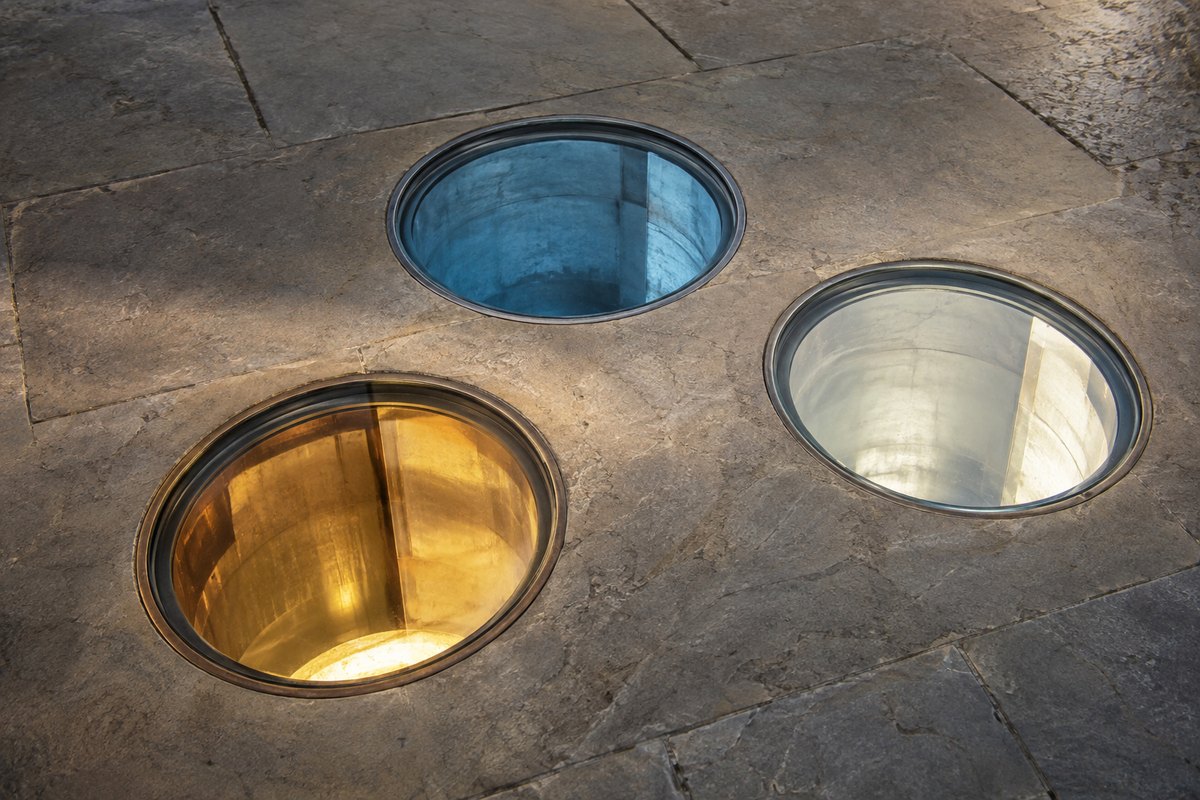 Drei unbeschriftete Lichtbrunnen im Steinboden, messingfarben, schieferblau und hell, ohne Zeichen